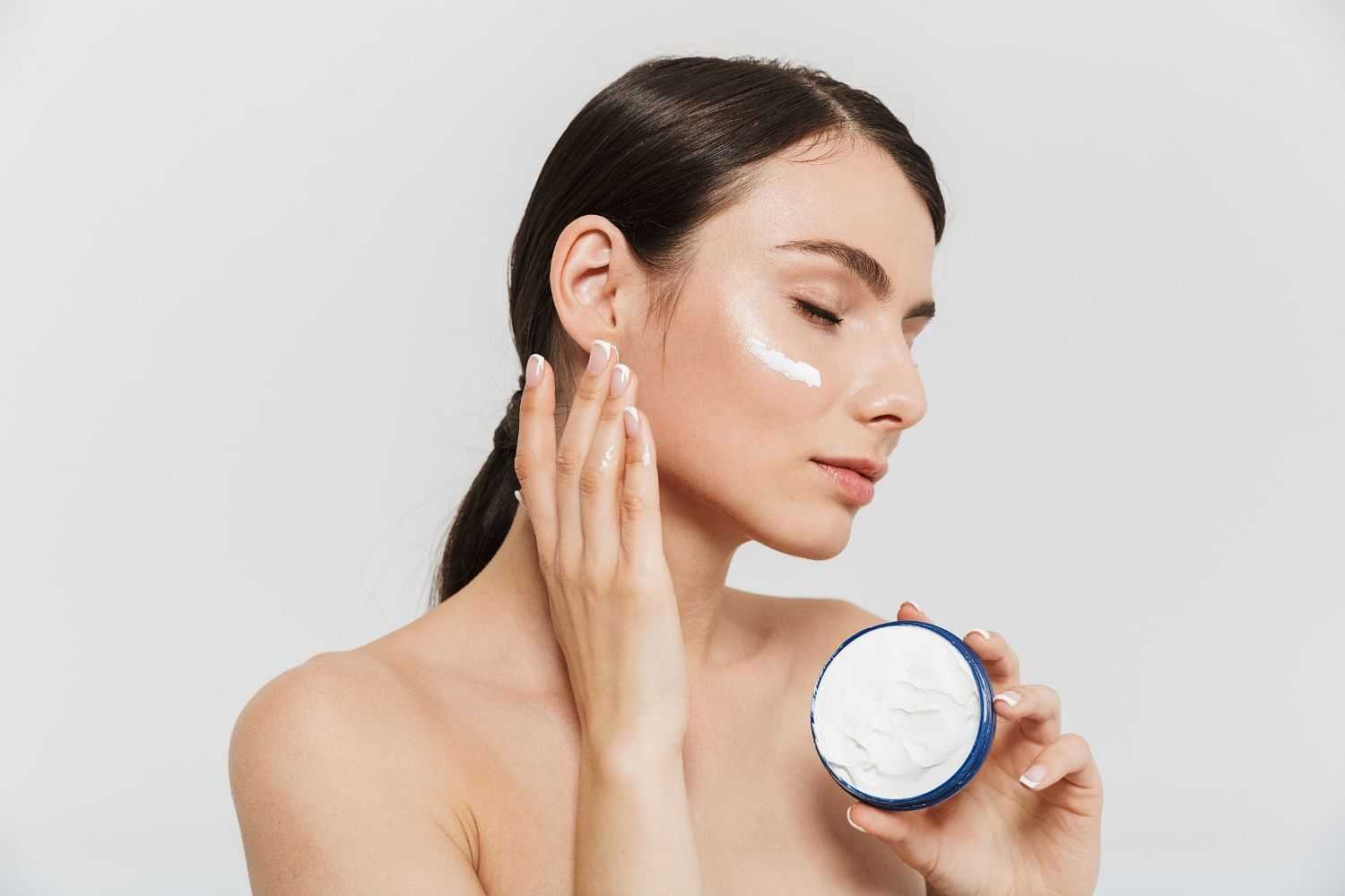 Woman applying facial cream on her cheek from a blue container with closed eyes.