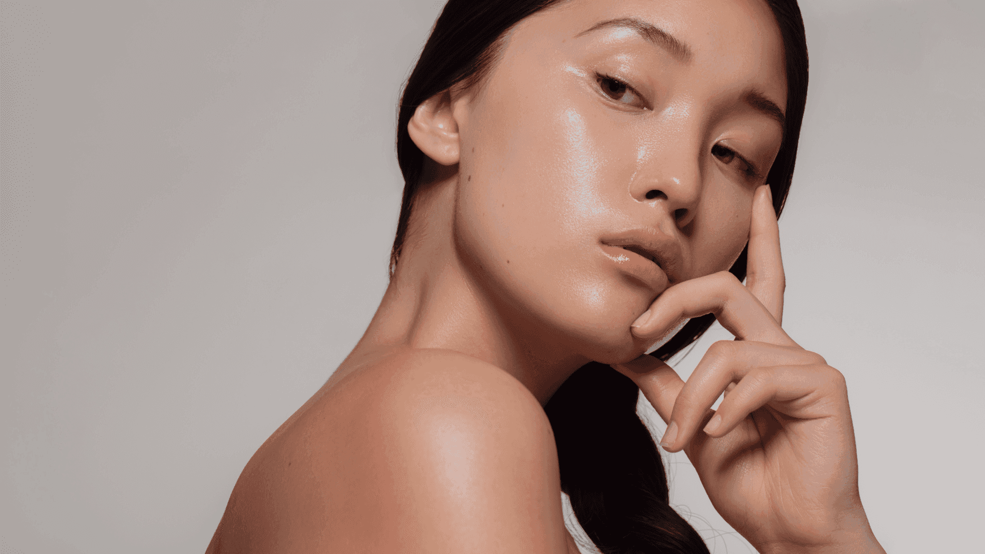 Woman with clear, glowing skin posing against a neutral background, touching her face.