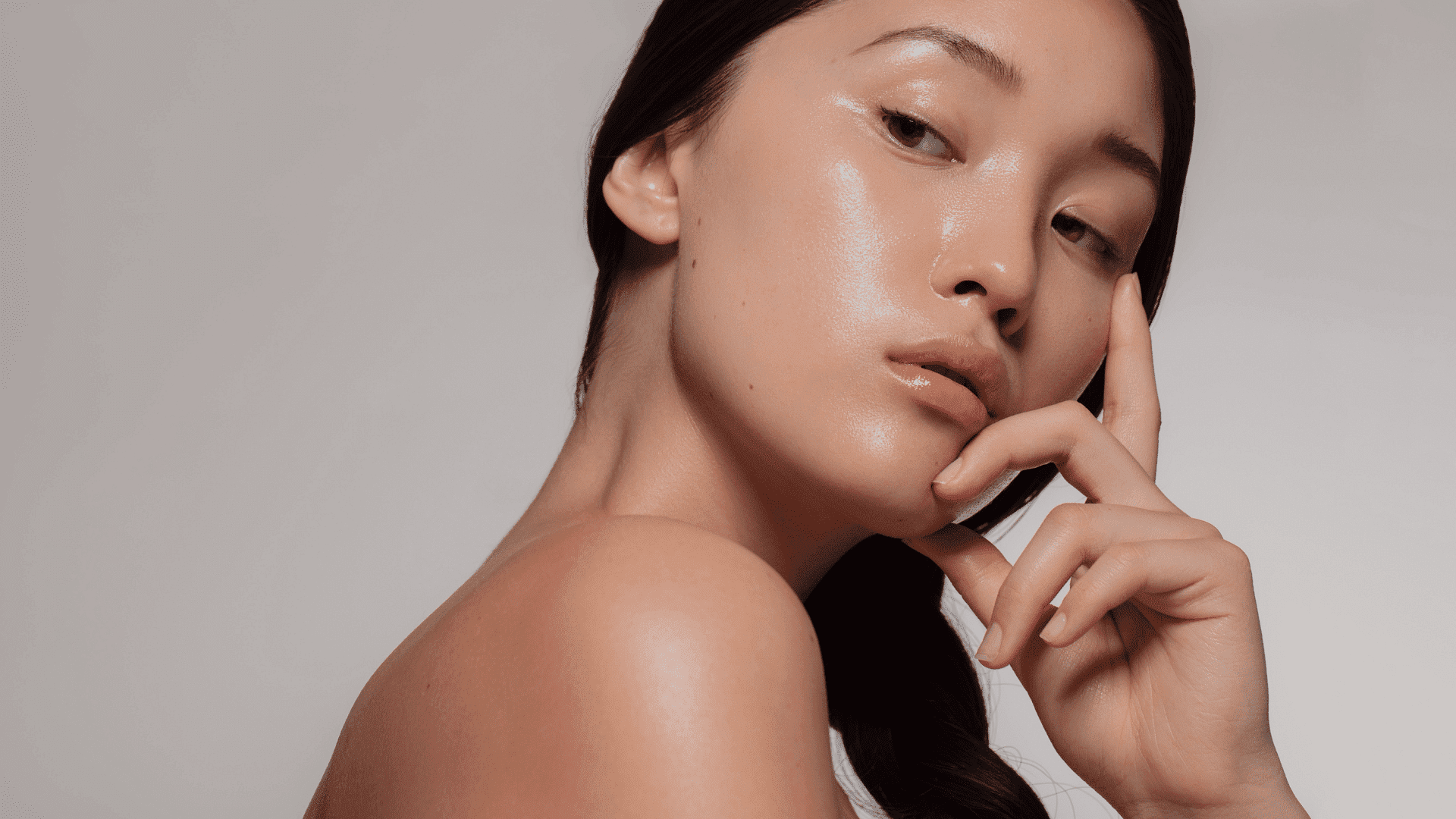 Woman with clear, glowing skin posing against a neutral background, touching her face.